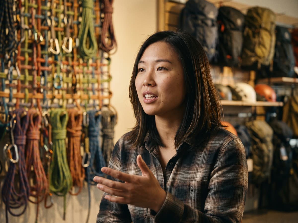 An Asian-Australian woman in her late 20s mid-sentence, in an outdoor-gear office with a climbing-rope wall decor
